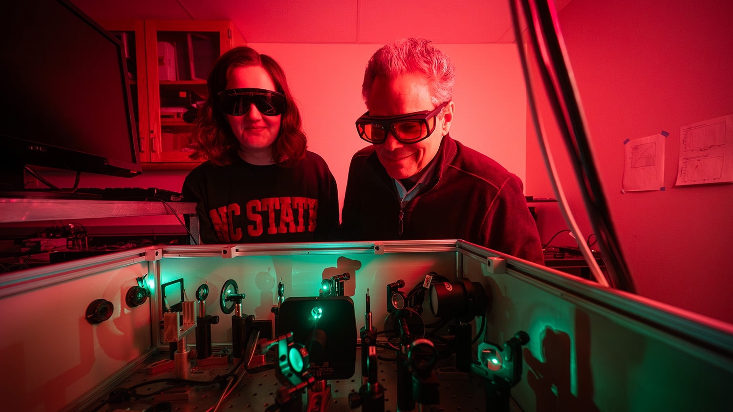 A student and a faculty member wear protective glasses as they look down into a box full of machinery, lenses and lights.